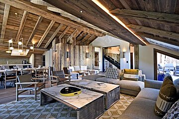 Luxurious chalet living room with exposed wood ceilings, plush sofas, marble coffee tables, and a dining area in the background.