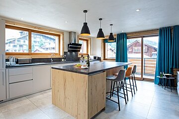 Modern kitchen with a wooden island, bar stools, black pendant lights, and windows looking out to alpine chalets.