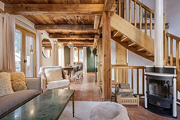 Cozy chalet interior featuring exposed wooden beams, a wooden staircase, a fireplace stove, and comfortable seating.