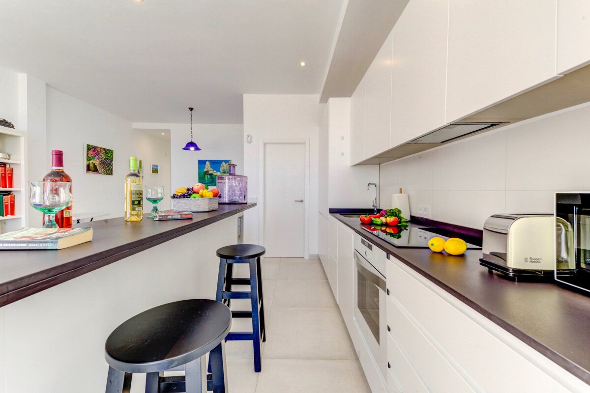 A bright, modern kitchen featuring a dark bar counter with stools. White cabinets, fresh produce, and wine create a sleek, inviting space.