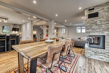Spacious open-plan living area with a large wooden dining table, a cozy stone fireplace, a modern kitchen, and a comfortable lounge section.