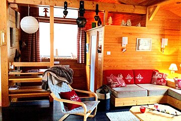 Cozy wooden chalet living room featuring a red sofa with festive pillows, a stylish armchair with a throw, and warm, rustic lighting.
