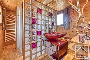 Warm wooden bathroom with a striking glass block wall (some magenta) separating the shower. Features twin red vessel sinks and a window.