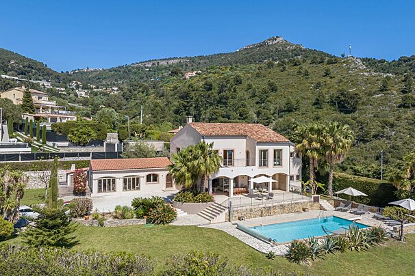 Mediterranean villa with a blue pool and patio, nestled among lush green mountains under a bright sky.