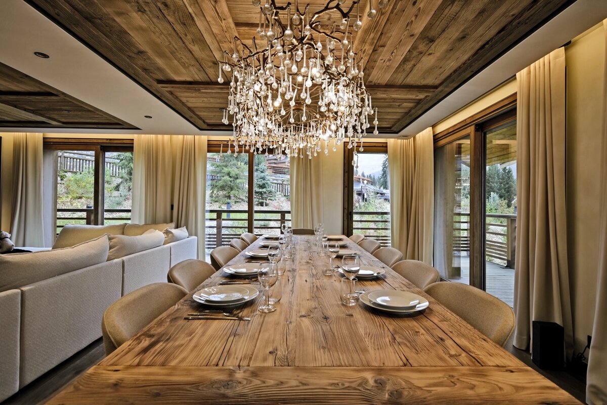 A luxurious rustic dining and living room with a long wooden table, ornate droplet chandelier, and large windows revealing a mountain landscape.