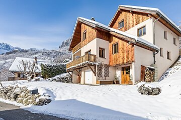 A wooden and stucco chalet on a snowy hillside, set against a background of mountains under a clear blue sky.