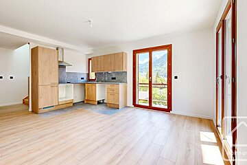 A bright, modern empty room with light wood flooring, kitchen cabinets, and a large window overlooking a scenic mountain view.