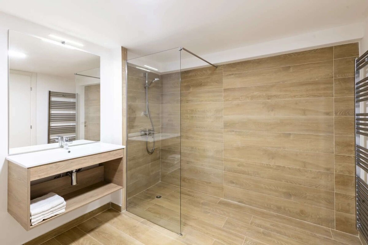 A modern bathroom featuring wood-look tiles, a glass shower enclosure, a white vanity with a large mirror, and a towel warmer.
