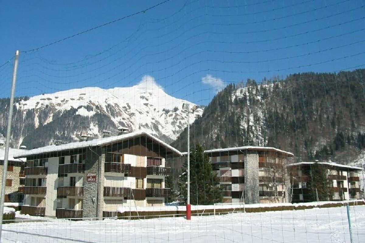 A large, multi-story wooden chalet with balconies, nestled against a rocky mountain backdrop. Pine trees and patches of snow complete the rustic scene.