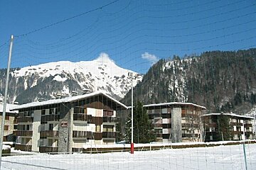 Snowy chalets with majestic snow-capped mountains in the background, viewed through a netting fence on a sunny day.
