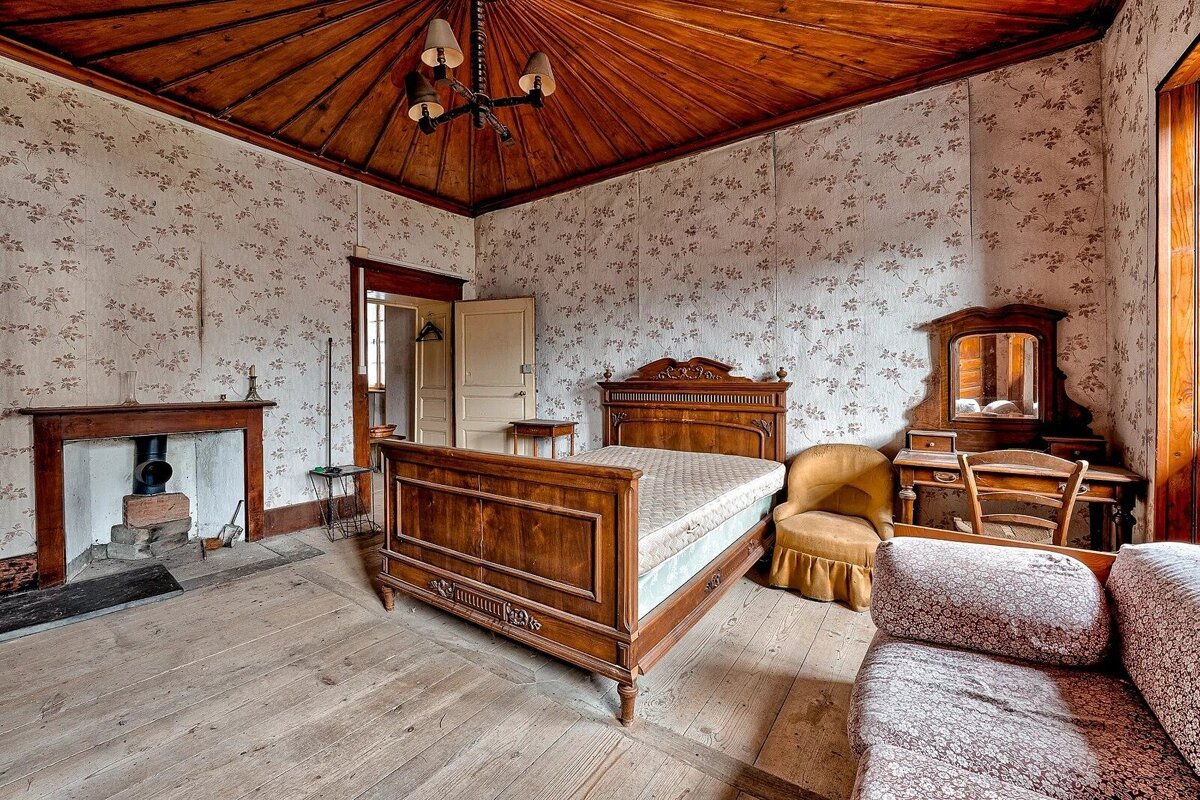 A vintage bedroom featuring patterned wallpaper, a carved wooden bed, dresser, fireplace, and a striking vaulted wooden ceiling with a chandelier.