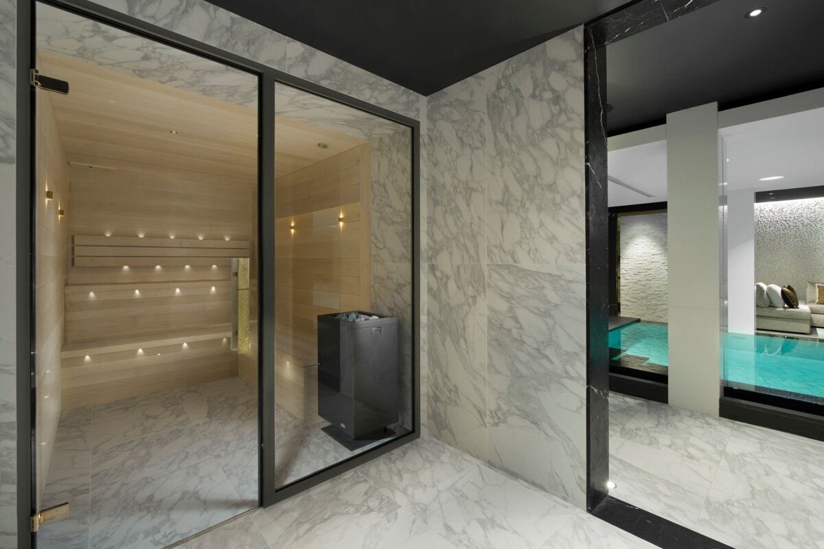 A modern spa area featuring a glass-front wooden sauna, black heater, white marble walls and floor, and a glimpse of a turquoise indoor pool.