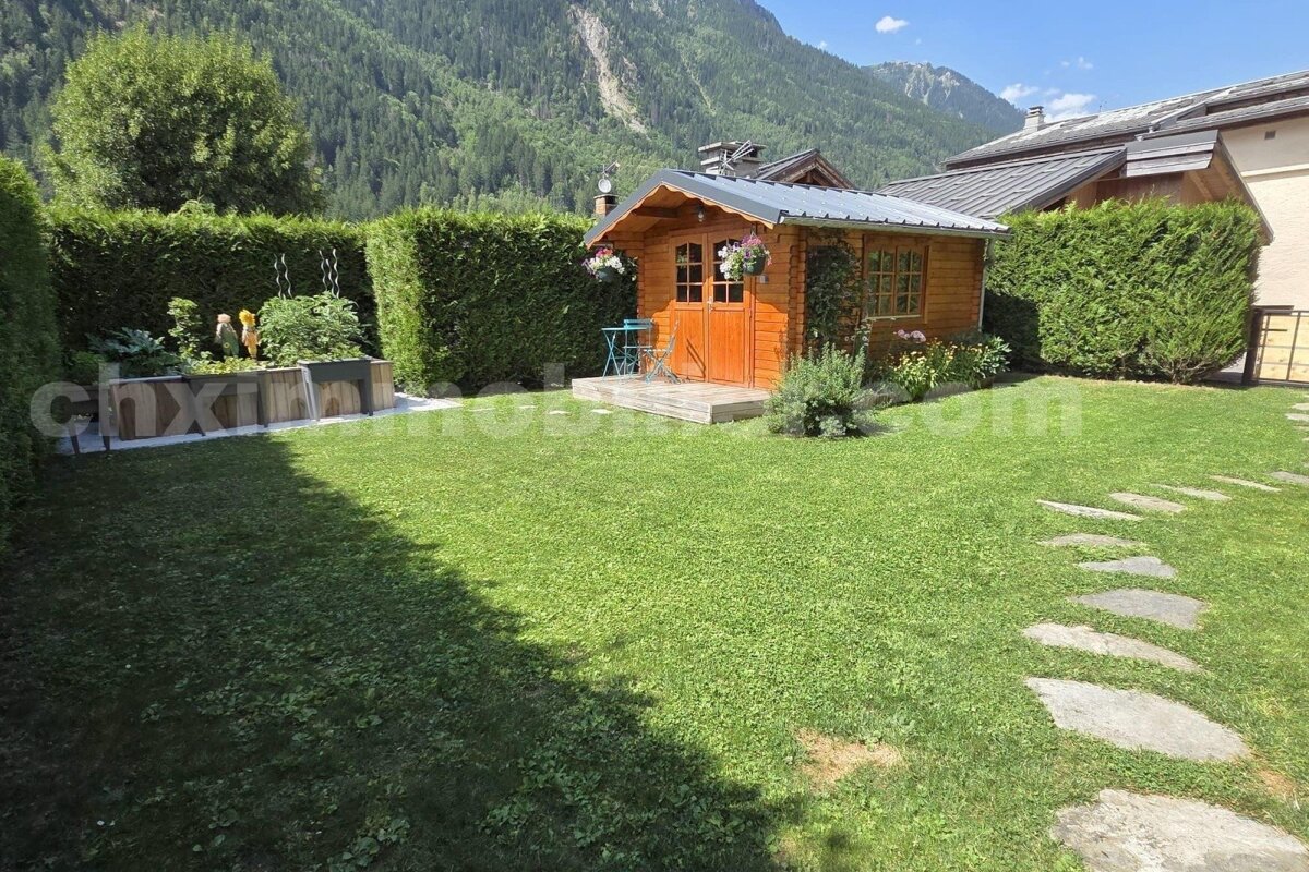 A charming wooden shed sits in a lush green garden with hedges, planters, and a stone path, all set against a backdrop of majestic mountains.