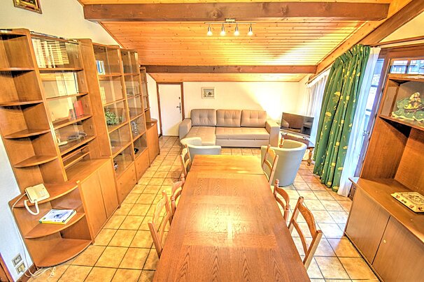 Cozy living-dining area with a long wooden table, tiled floor, large bookshelves, and a beige sofa under a rustic wood ceiling.