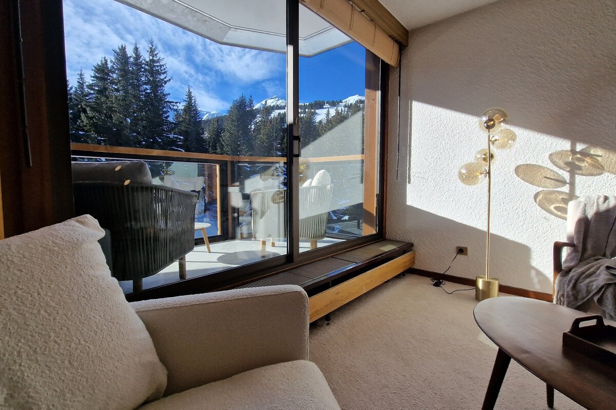 A cozy room with a large window and balcony overlooking a snowy mountain and pine forest. Sunlight streams in, illuminating the comfy interior.