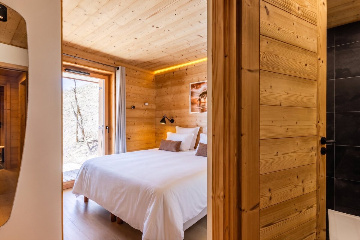 Cozy wooden chalet bedroom with a white bed, wood-paneled walls, and an open door showing a winter landscape.