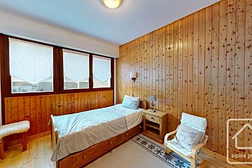 Cozy, rustic bedroom with wood-paneled walls, a single bed, windows with white blinds, a nightstand, and a light chair.