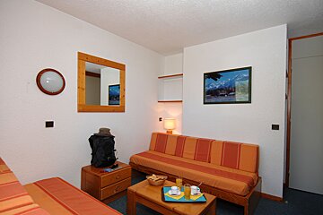 A small, functional studio room featuring an orange striped sofa bed, a single bed, and a coffee table with breakfast. Wall decor includes a mirror and mountain art.