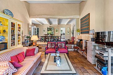 A cozy, rustic living room with a plaid sofa, red armchairs, fireplace, and exposed beams. Terracotta tiles lead to an elevated dining room.