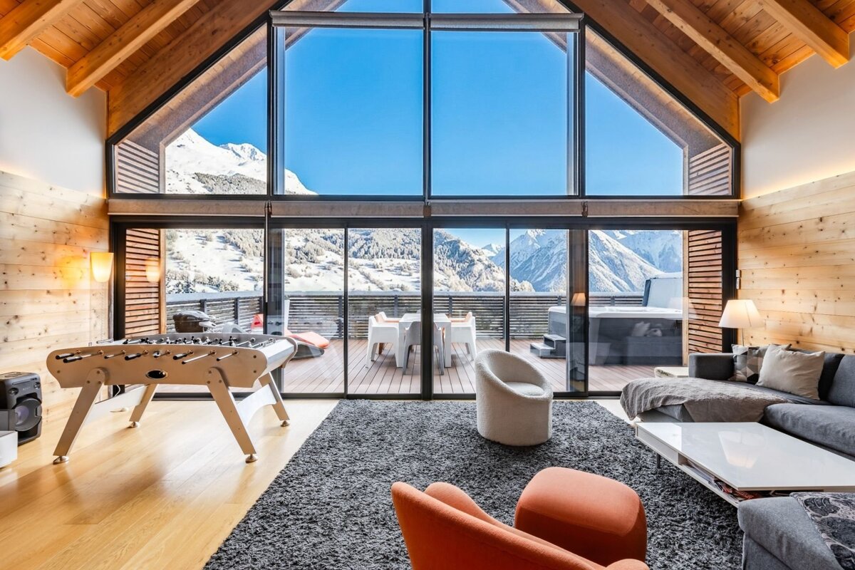 Modern chalet interior featuring a foosball table, cozy seating, and massive windows overlooking a snowy mountain landscape.
