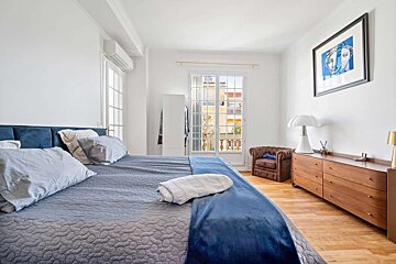 A bright, modern bedroom with a blue bed, large window to a balcony, wooden floor, brown armchair, dresser, and abstract art.