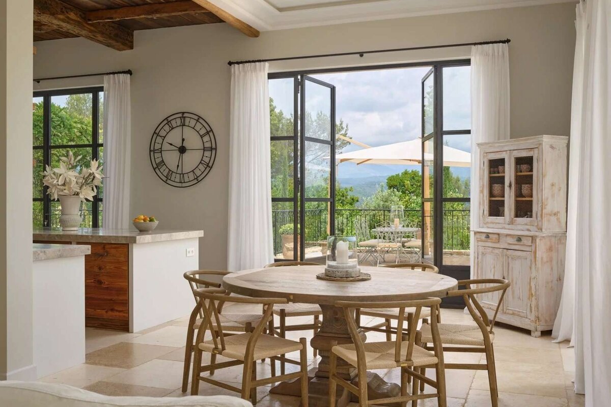 A rustic dining room features a round wooden table, woven chairs, and large glass doors opening to a scenic balcony overlooking green hills.