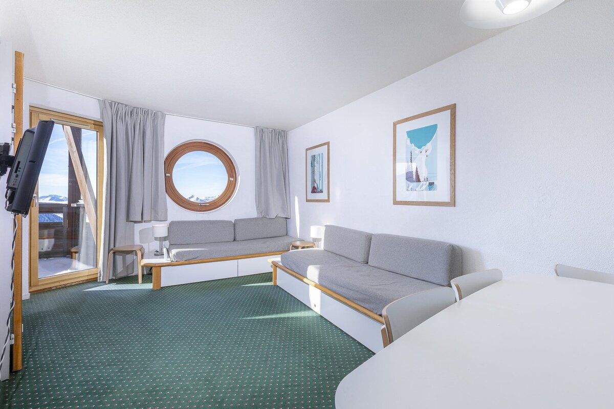 A bright living room with white walls, green carpet, two sofas, a round window & balcony overlooking snow-capped mountains.