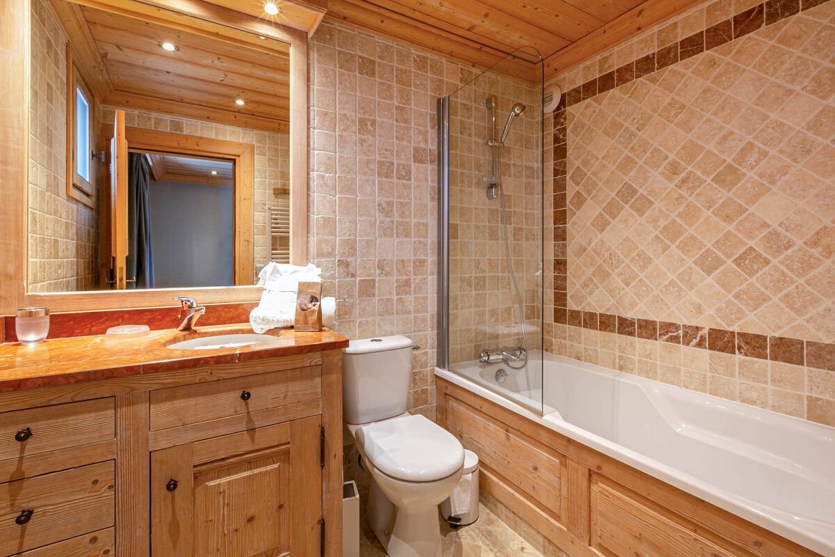A warm bathroom featuring a wooden ceiling and vanity, beige/brown tiled walls, a bathtub with glass shower screen, toilet, and sink.