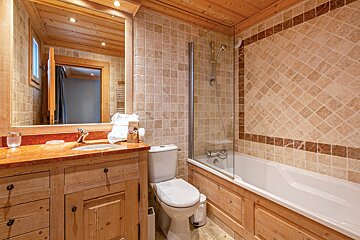 A warm bathroom featuring a wooden ceiling and vanity, beige/brown tiled walls, a bathtub with glass shower screen, toilet, and sink.