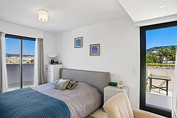 Bright, modern bedroom featuring a grey bed with a blue throw blanket and glass doors opening to sunny balconies with views.