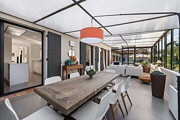 Modern sunroom with a wooden dining table, white chairs, and a lounge area under a translucent roof, offering outdoor views.