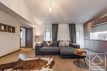 A modern living space featuring a grey sectional sofa, cowhide rug, wooden floors, and a small kitchen corner.