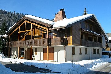 A wooden alpine chalet with a snow-covered roof and balconies, set in a snowy winter landscape under a clear blue sky.