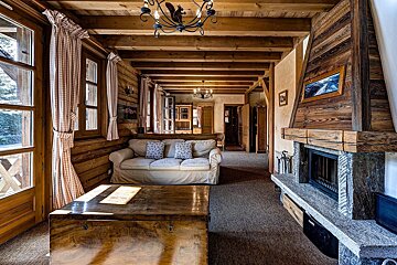 Cozy, wood-paneled rustic living room with exposed beams, stone fireplace, sofa, and large windows. A squirrel chandelier hangs overhead.