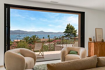 A modern living room with large sliding doors opening to a scenic balcony overlooking a bay with boats and mountains.