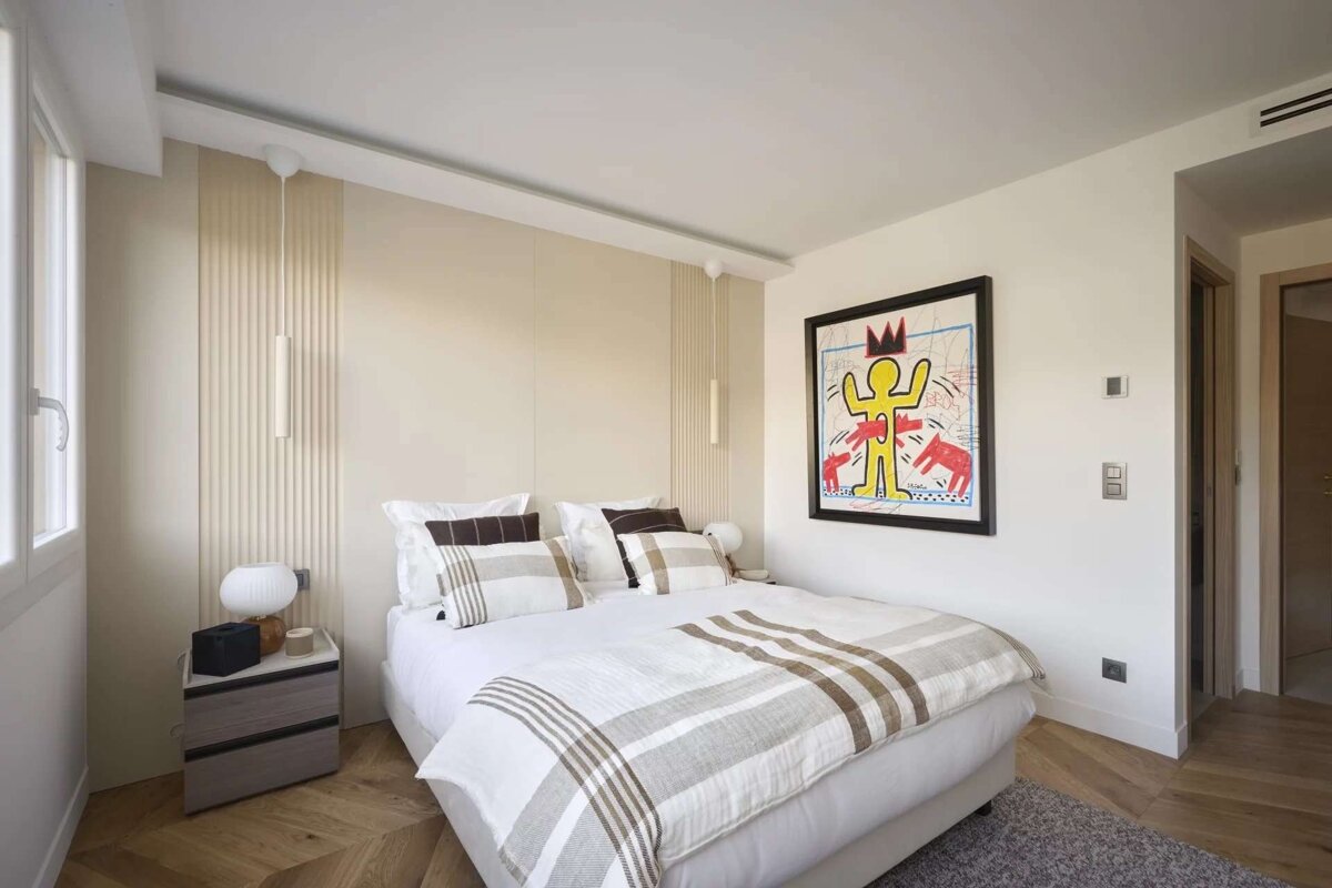 A modern bedroom features a large bed with white and striped bedding, a vibrant Keith Haring artwork, and light-colored walls. Wood floors complete the look.
