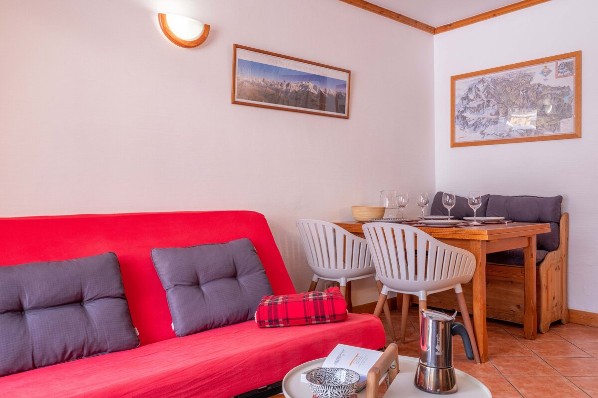 A cozy living/dining area featuring a bright red sofa, wooden dining table with white chairs & bench, and mountain-themed wall art.