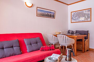 A cozy living/dining area featuring a bright red sofa, wooden dining table with white chairs & bench, and mountain-themed wall art.