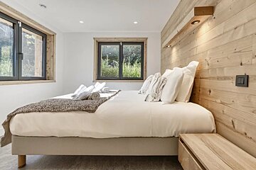 Cozy, modern bedroom featuring a bed with white linens, a light wood-paneled accent wall, and windows overlooking greenery.