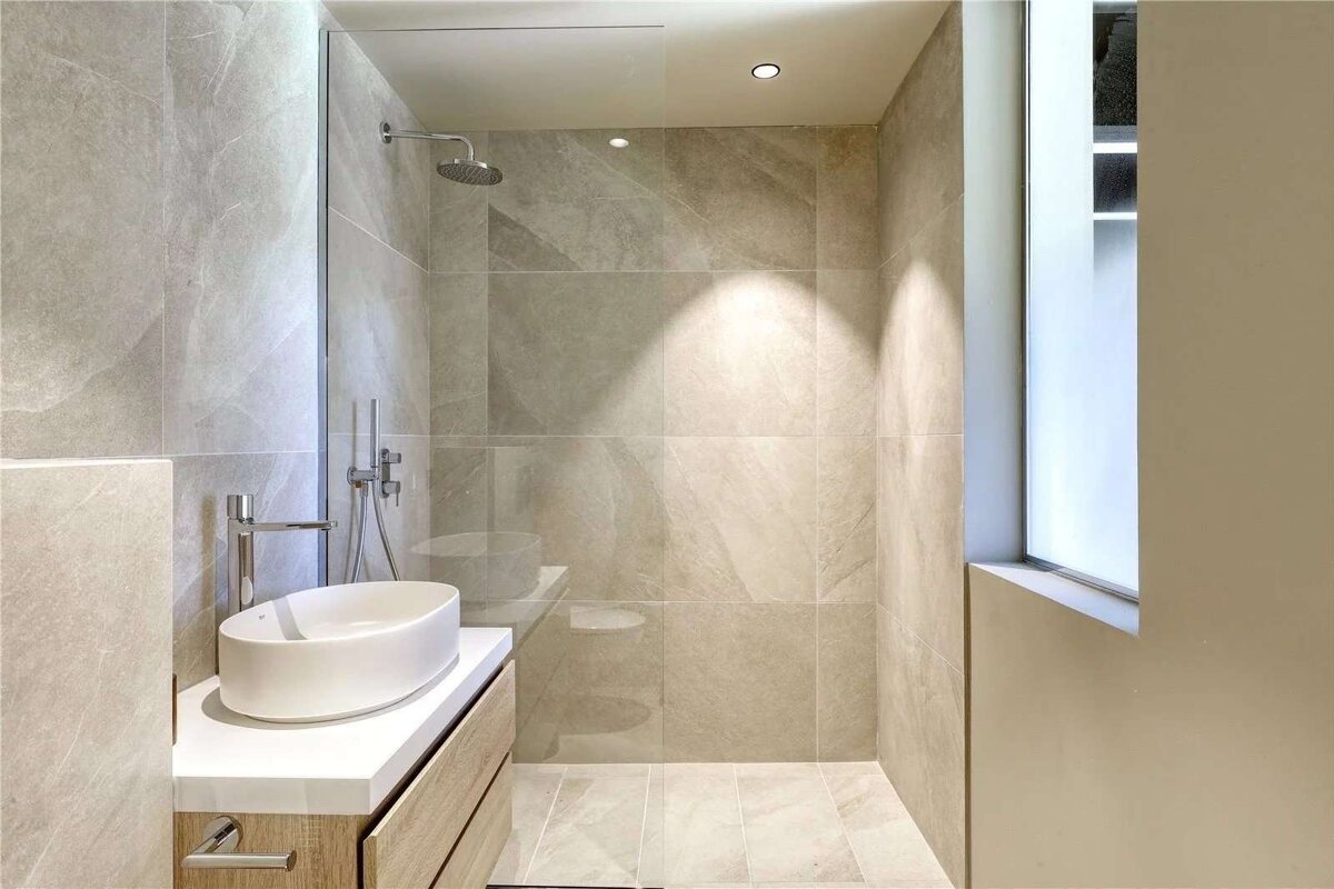 Modern bathroom with neutral tiled walls, walk-in shower, vessel sink on wooden vanity, recessed lighting, and frosted window.