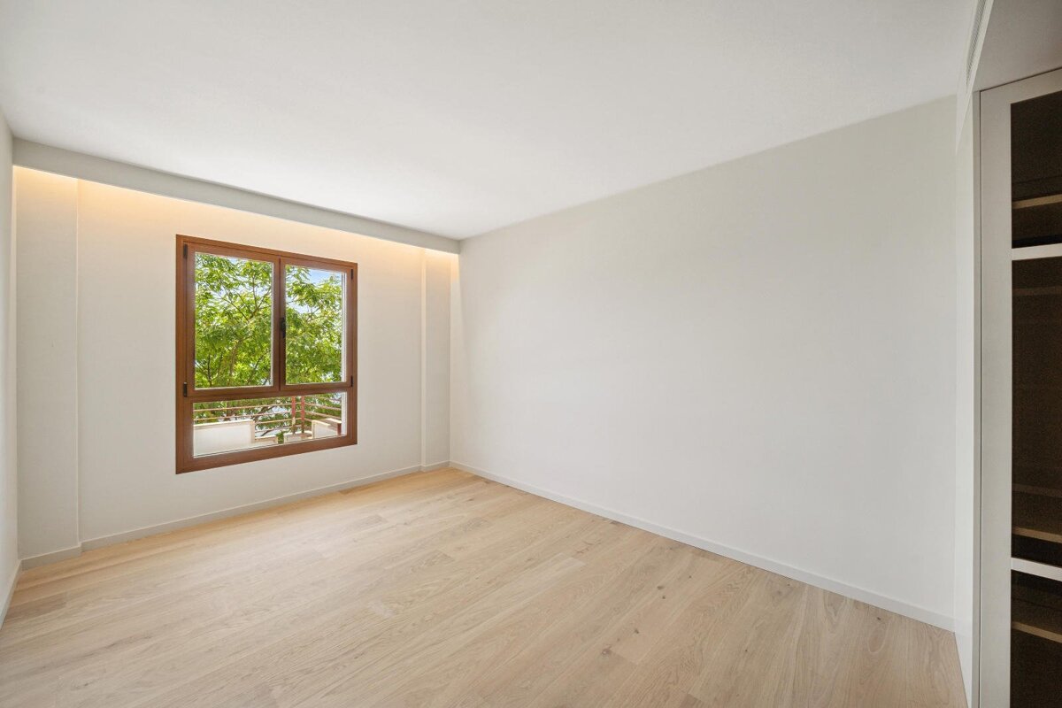 An empty room with white walls, light wood floor, a window framing green trees, and subtle ceiling lighting. A built-in is on the right.