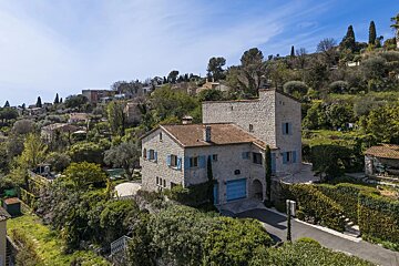 A stone villa with blue shutters, nestled in a lush green hillside under a clear blue sky.