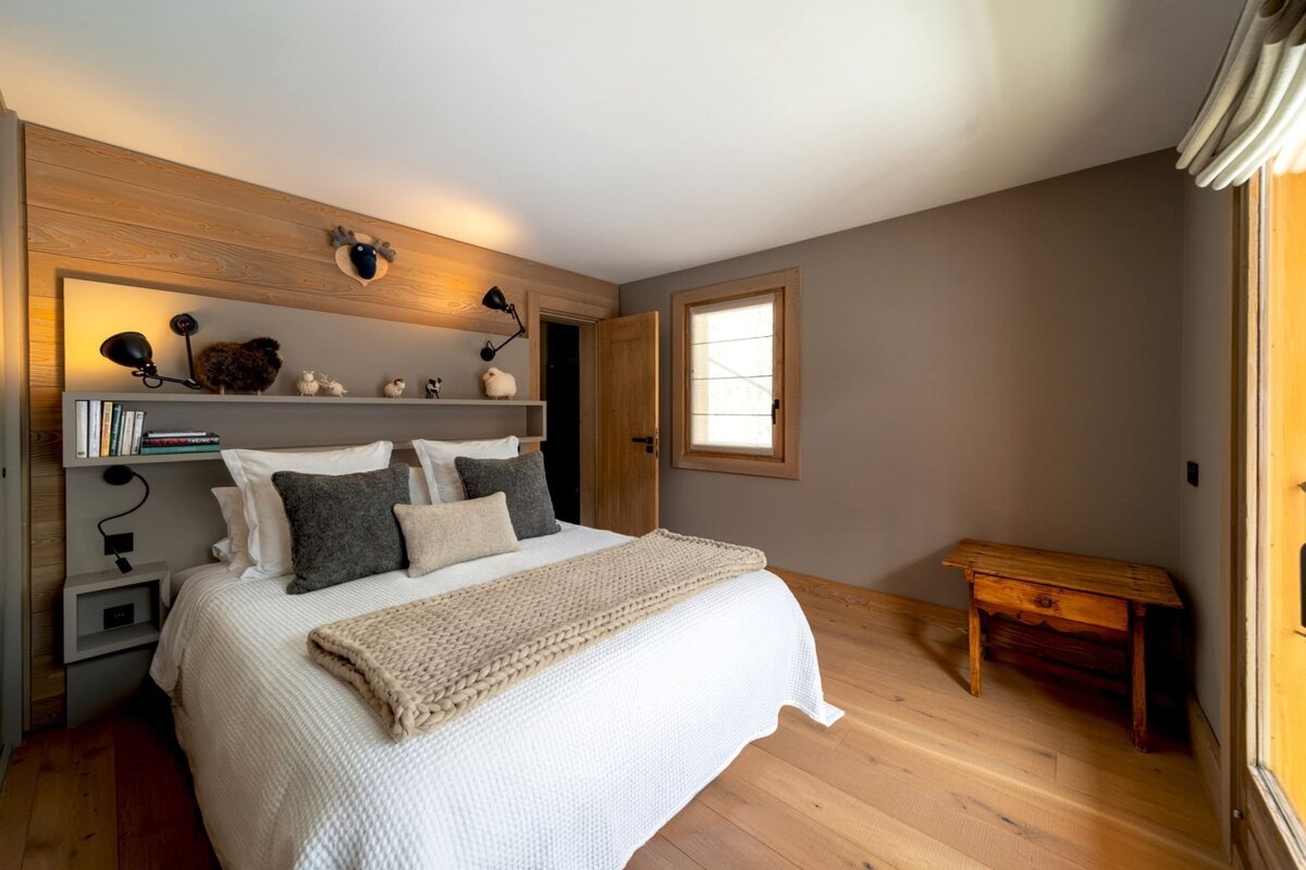 Cozy, rustic-modern bedroom with a large bed, wooden walls and floor. A shelf above the bed holds books and sheep-themed decor.