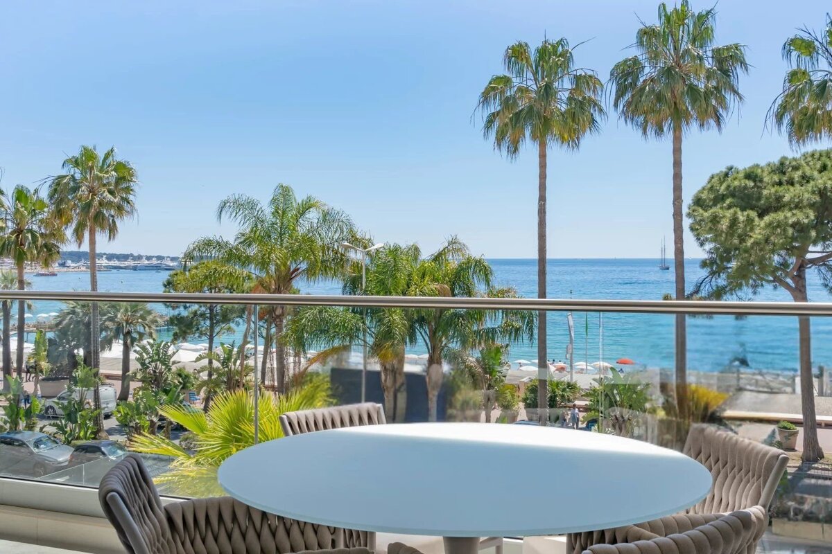 Sunny balcony view of a blue ocean, sandy beach with umbrellas, and lush palm trees. A white round table and chairs are in the foreground.