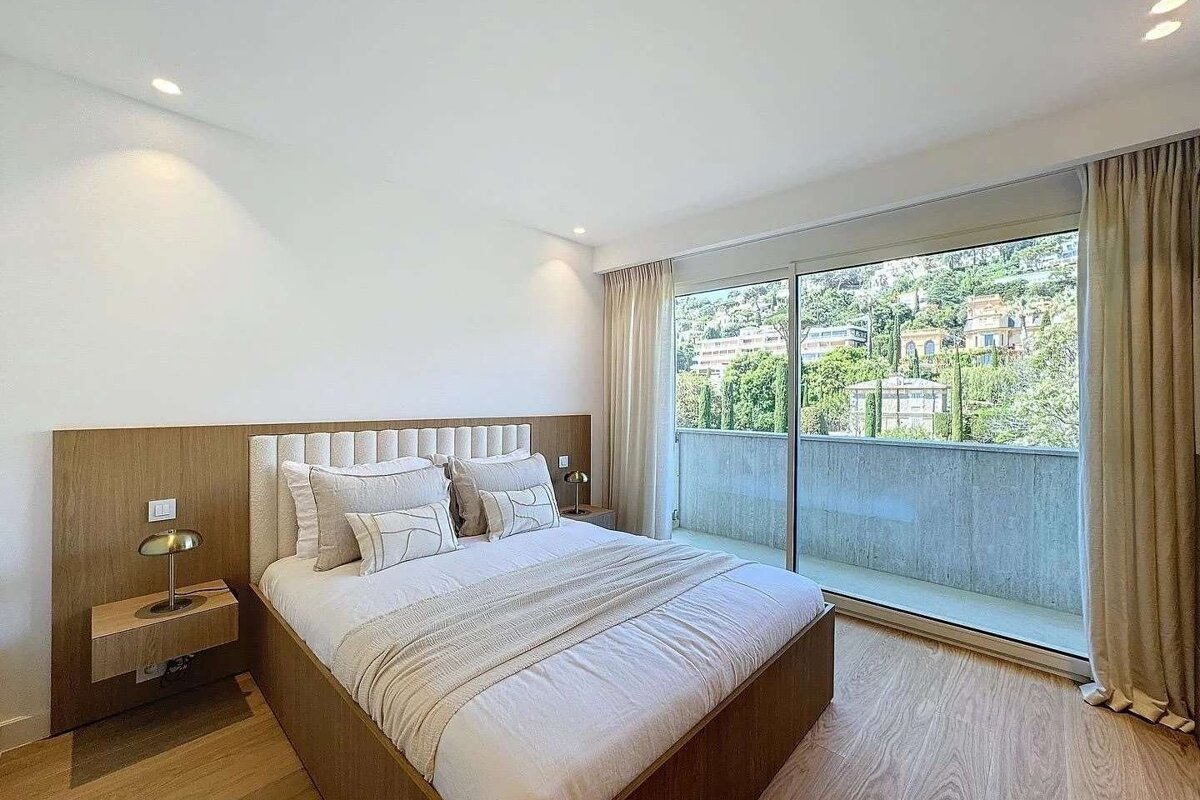 A bright, modern bedroom features a large bed, wood details, and a floor-to-ceiling glass door offering a panoramic view of lush, hilly landscape.