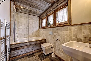 Rustic chalet-style bathroom with a wooden ceiling, tiled walls/floor, bathtub with shower screen, toilet, sink, and windows.