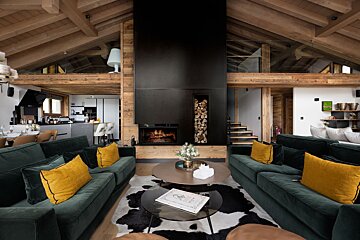 Chic chalet living room featuring a grand fireplace, exposed wooden beams, dark green sofas with yellow pillows, and a cowhide rug.