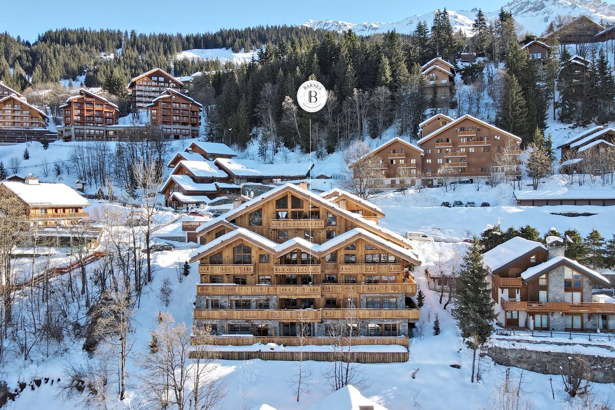 A large, beautiful wooden chalet in a snow-covered mountain village, surrounded by other chalets and pine trees under a bright winter sky.