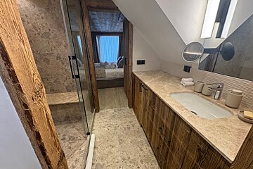 Modern rustic bathroom with a wooden vanity, stone countertop, glass shower, and a doorway leading into a bedroom.