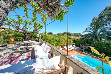 Bright patio with a vine pergola, dining area & lounge overlooking a blue pool with sun loungers & lush greenery under a clear sky.